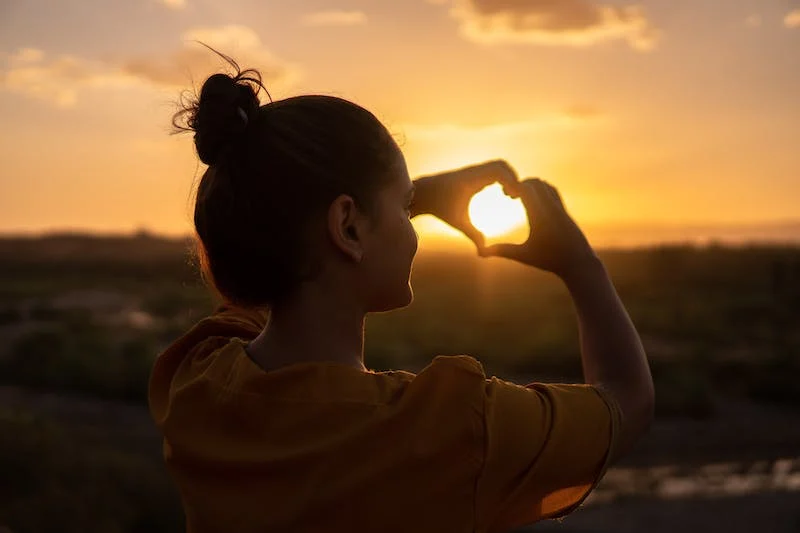 A girl making a heart symbol with her hands towards the sun
